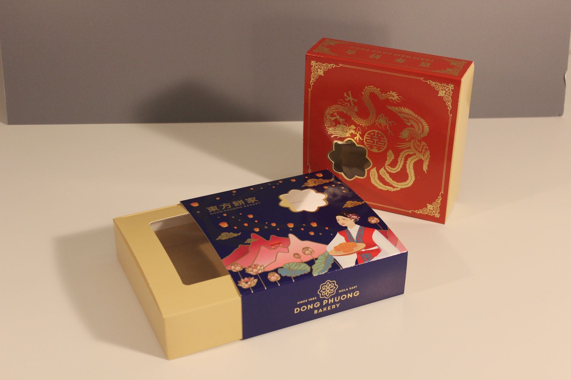 Two die cut Dong Phuong Bakery mooncake cartons, a red one printed with a gold dragon and a navy one with a die cut window
